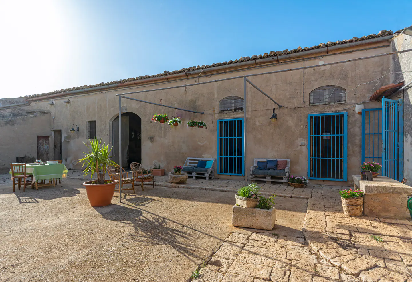 Outdoor courtyard with stone floor, rustic building, blue doors, seating areas, and potted plants under a clear blue sky.