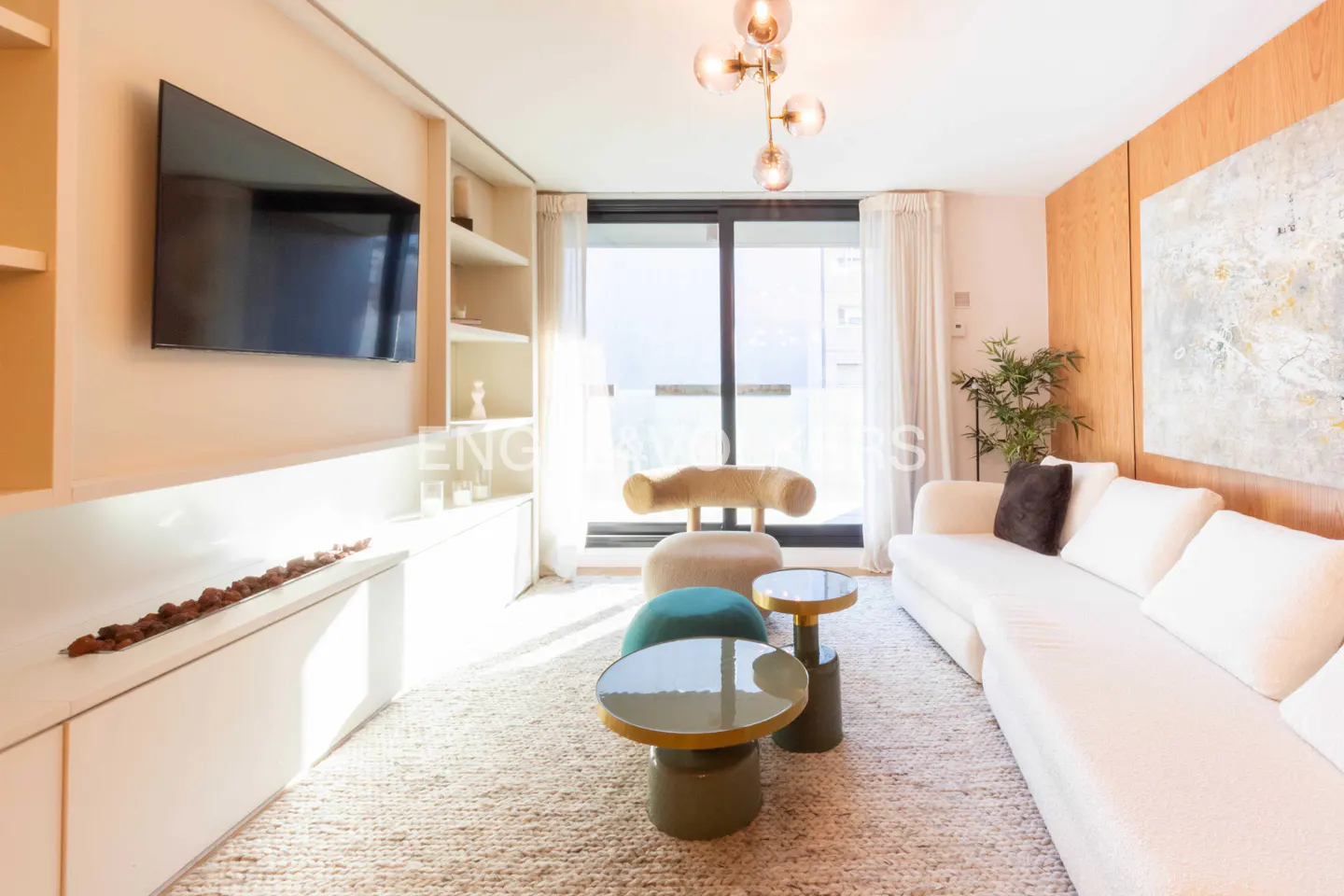 Bright living room with white sofa, beige chair, and green tables. A TV hangs above a white cabinet, and a modern chandelier illuminates the space.