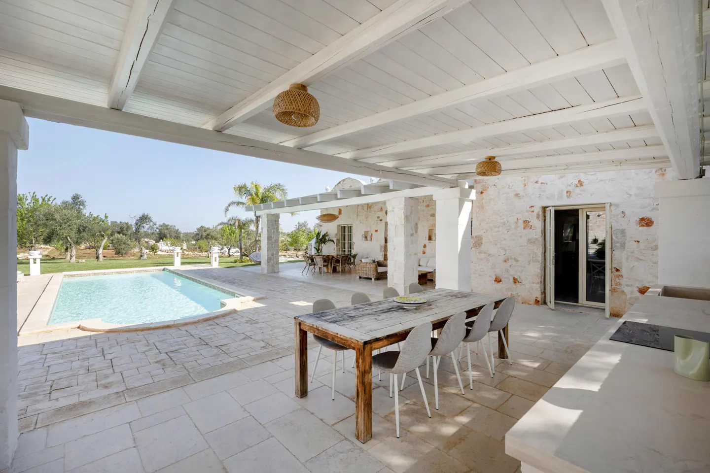 Outdoor patio with a pool. A long wooden table with gray chairs sits under a white pergola with wicker light fixtures. A pool and green lawn are visible.