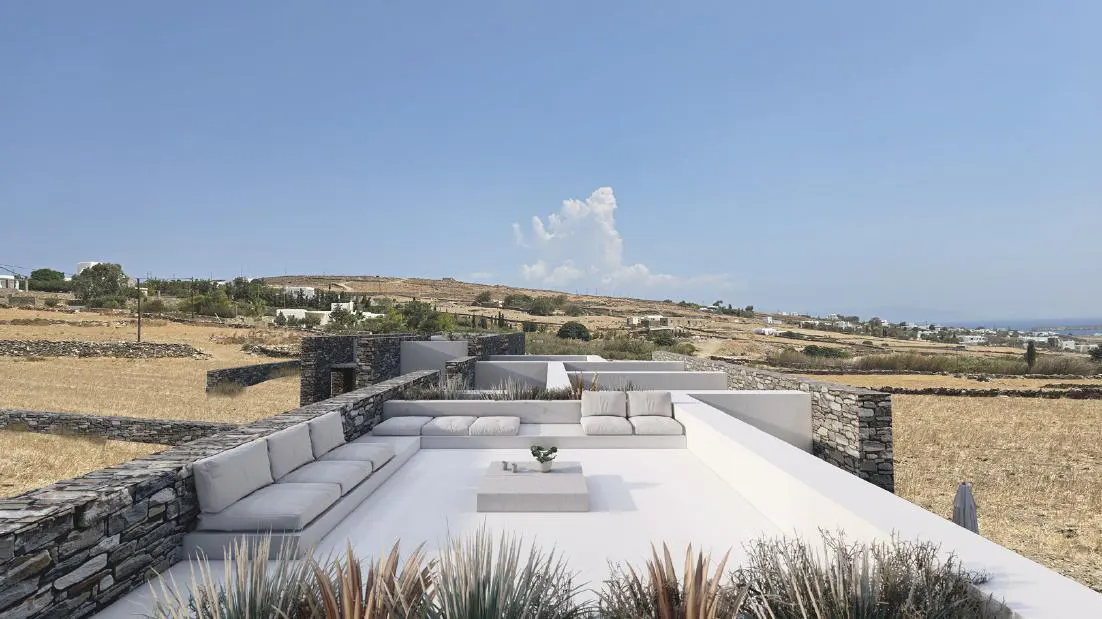Rooftop lounge area with white sofas and table, stone walls, and a view of a dry, grassy landscape under a blue sky.