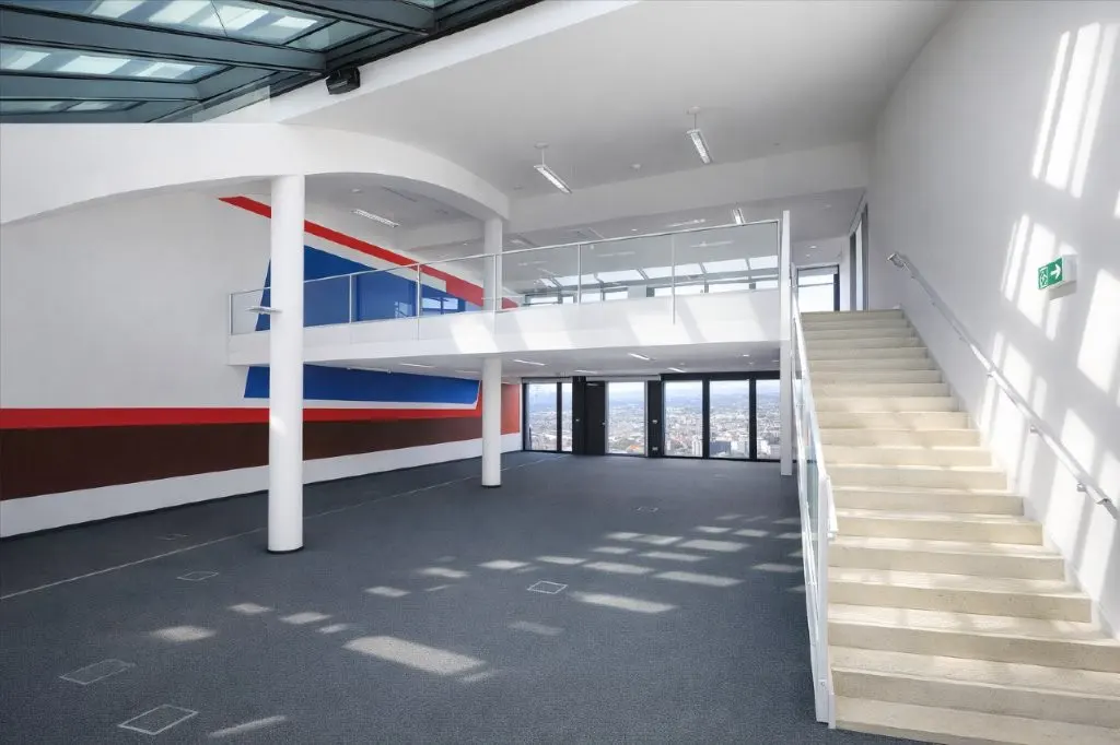 Bright, modern office space with gray carpet, white walls, and a staircase. A second-floor balcony overlooks the main area, with city views visible through windows.
