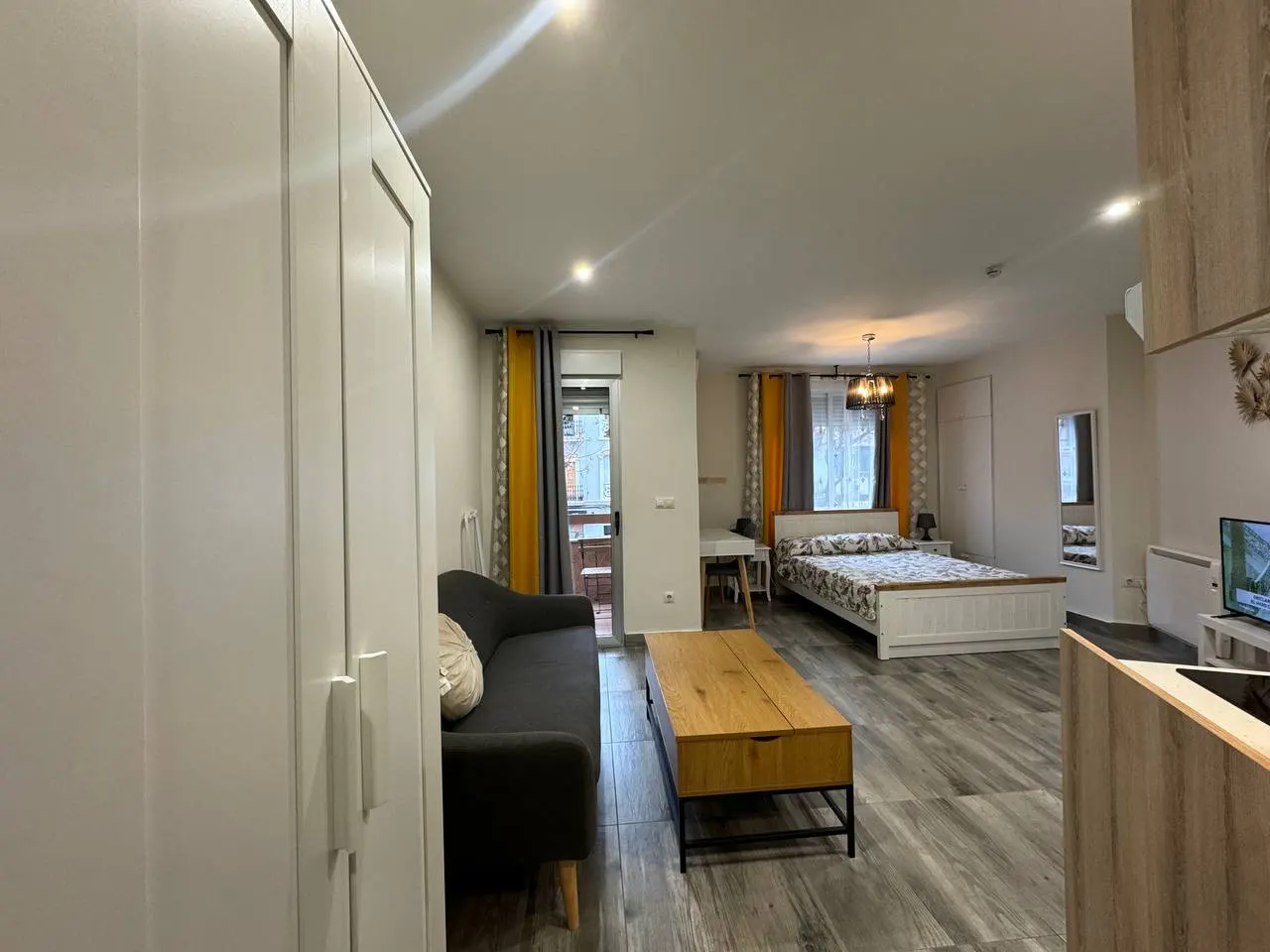 A studio apartment with a gray sofa, wooden coffee table, and white bed with floral bedding. Yellow and gray curtains frame the windows.