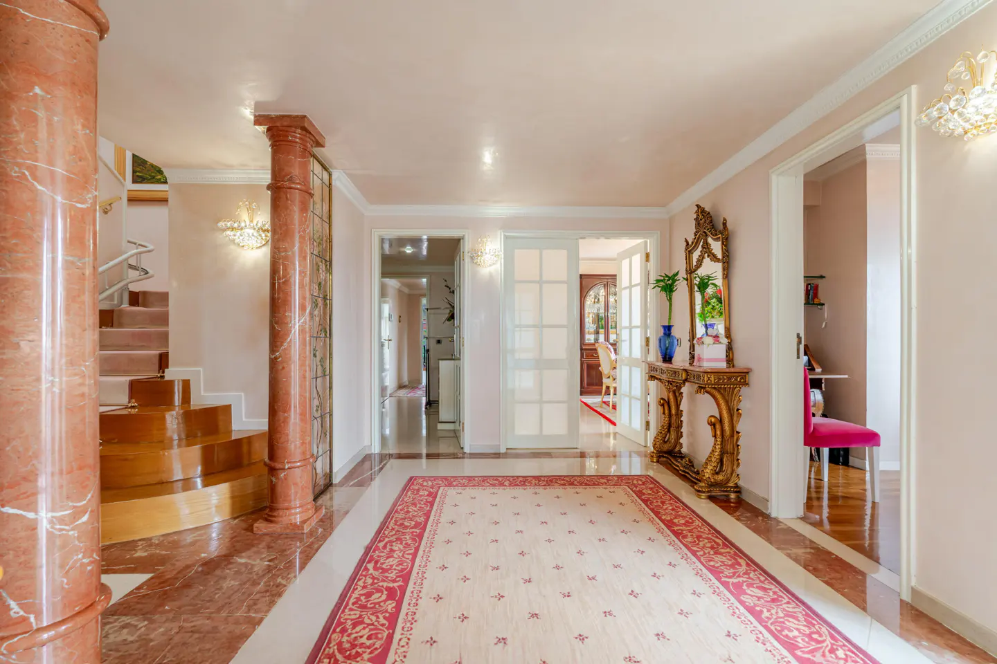 Elegant foyer with marble floors, columns, and a patterned rug. A gold console table with a mirror stands against a light pink wall.