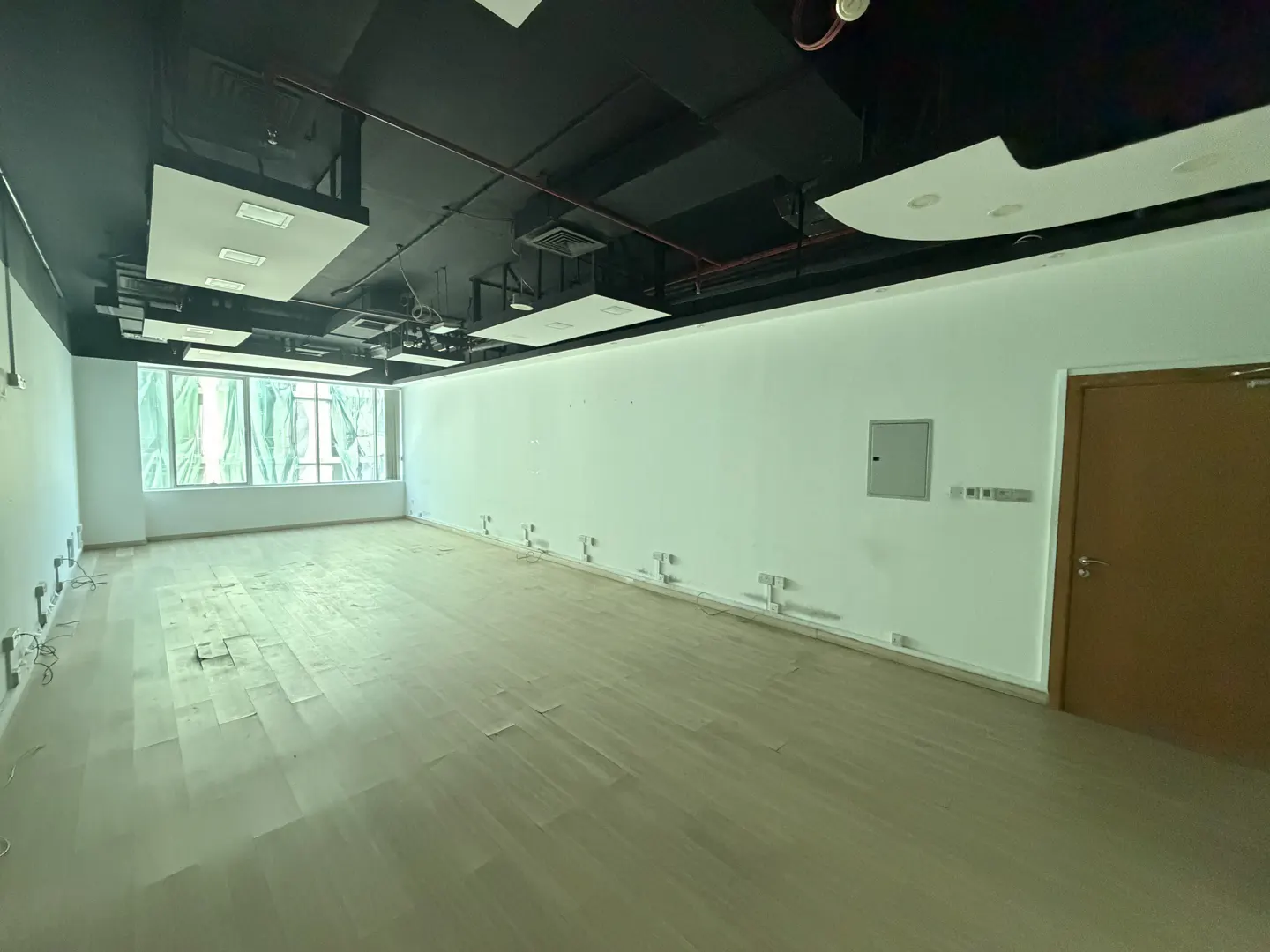 Empty office space with light wood floors, white walls, and a black ceiling with recessed lighting. A window with sheer curtains is on the left.