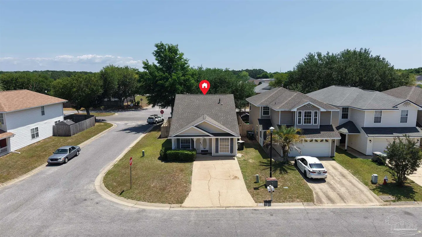 Aerial view of a light yellow single-family home with a gray roof and blue shutters in a suburban neighborhood. A red pin marks the house's location.