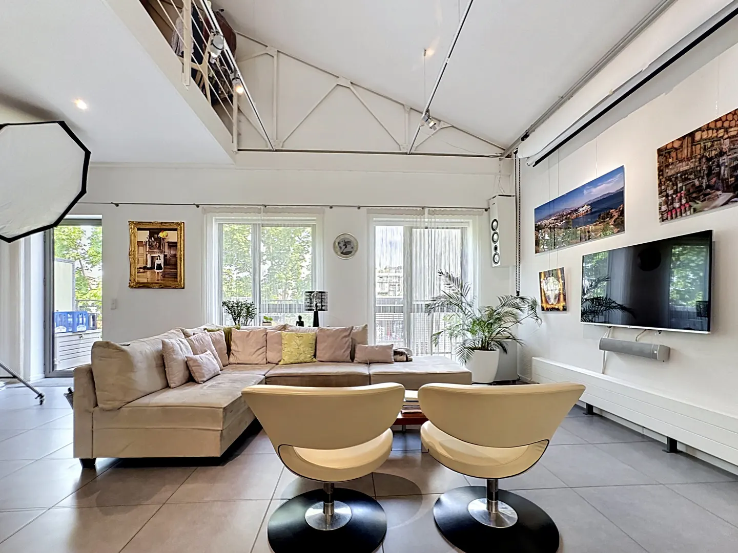 Bright living room with white walls, a beige sectional sofa, two beige chairs, and large windows. Art hangs on the wall.