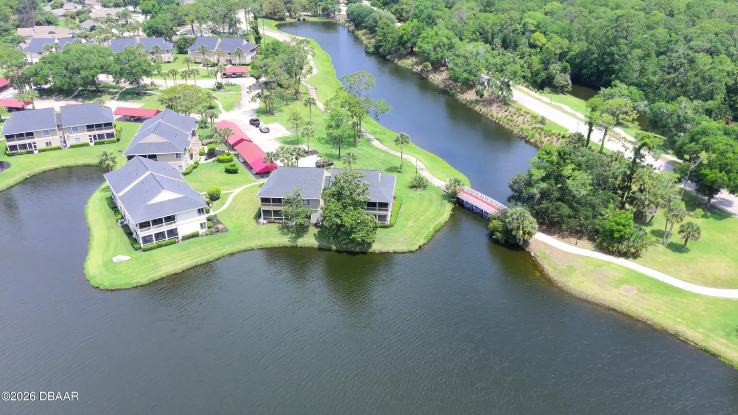 Aerial view of condos on a lake, with a red-roofed carport, a bridge, and green trees.