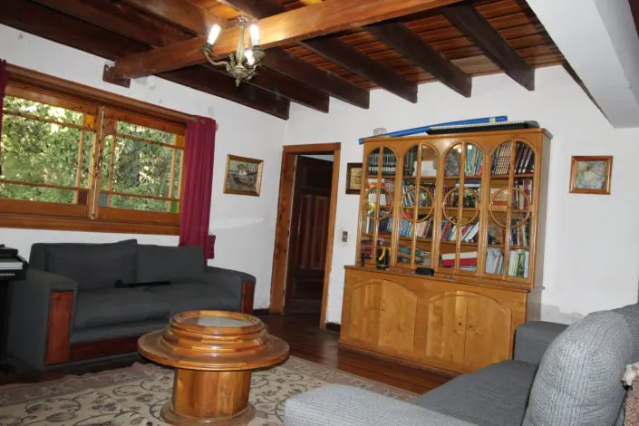 Living room with wood floors, exposed beam ceiling, and a large window. A gray sofa, round wood table, and bookcase are also visible.