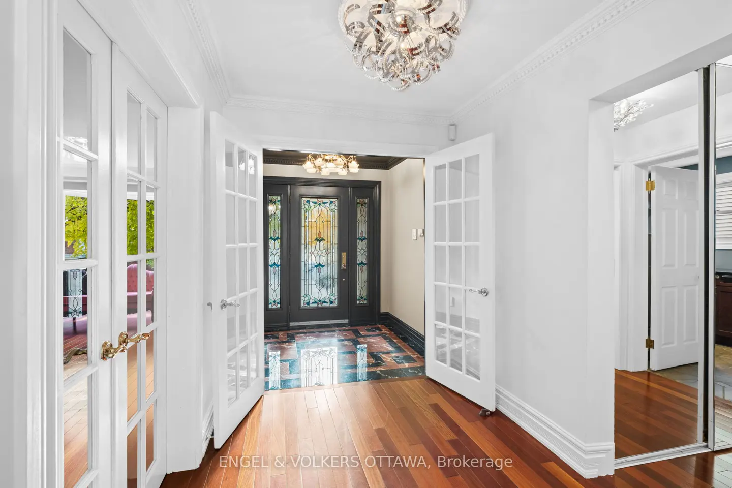 A bright foyer with hardwood floors, white walls, and a decorative glass front door. French doors lead to other rooms. A modern chandelier hangs from the ceiling.