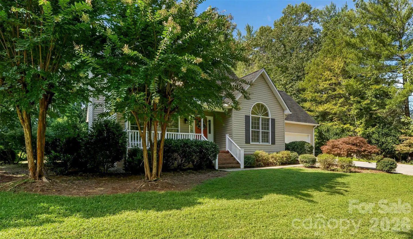 A one-story house with a porch, green lawn, and trees. The house is light gray with a dark roof and a red front door. "For Sale" sign on the lawn.