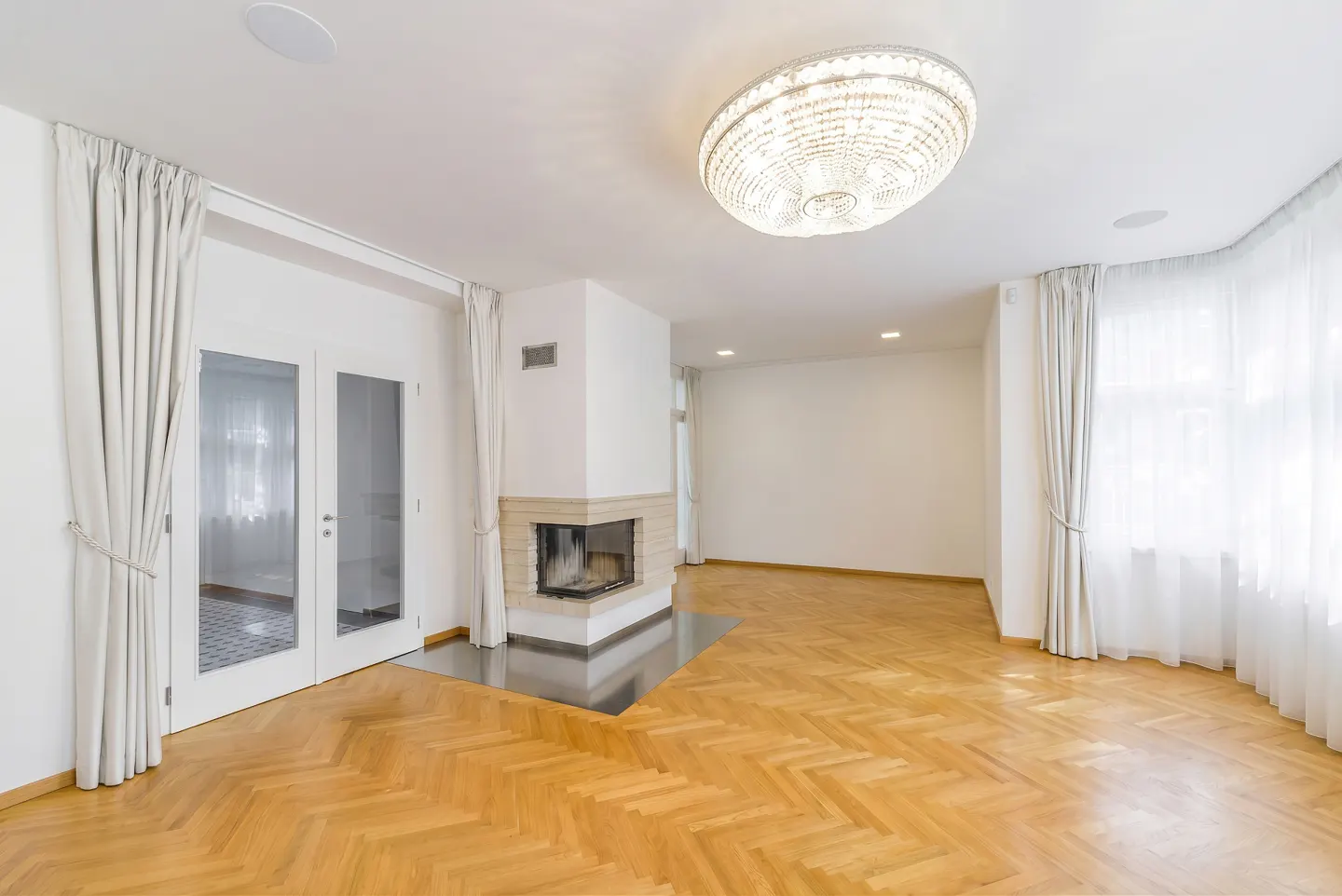Bright living room with herringbone wood floors, a corner fireplace, white walls, floor-to-ceiling curtains, and chandelier.