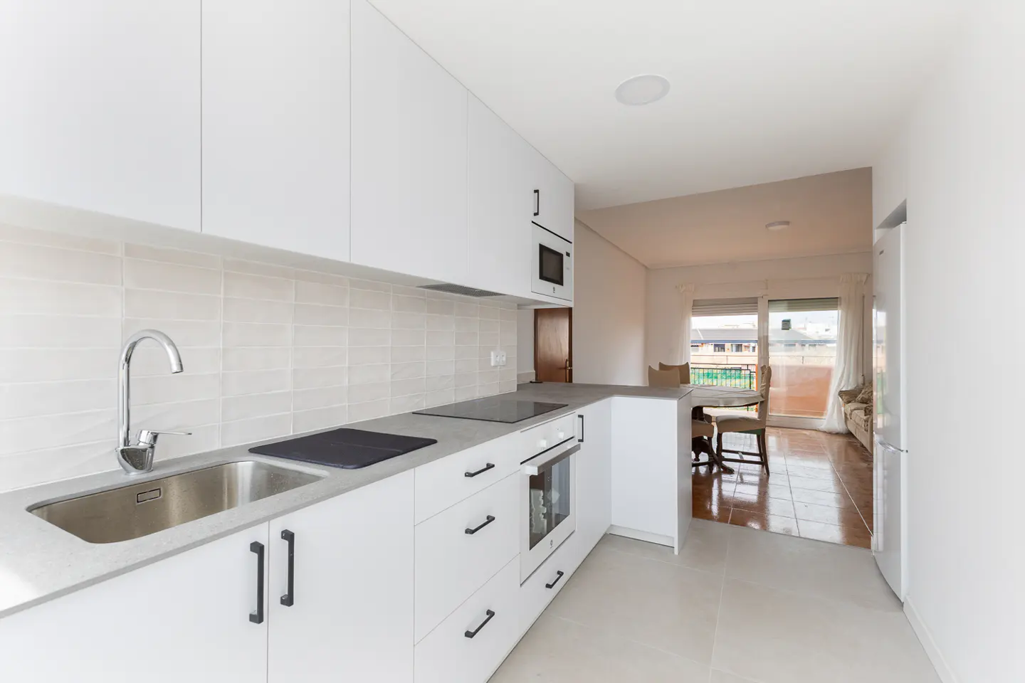 Bright, modern kitchen with white cabinets, gray countertops, and stainless steel sink. Dining area visible in the background.