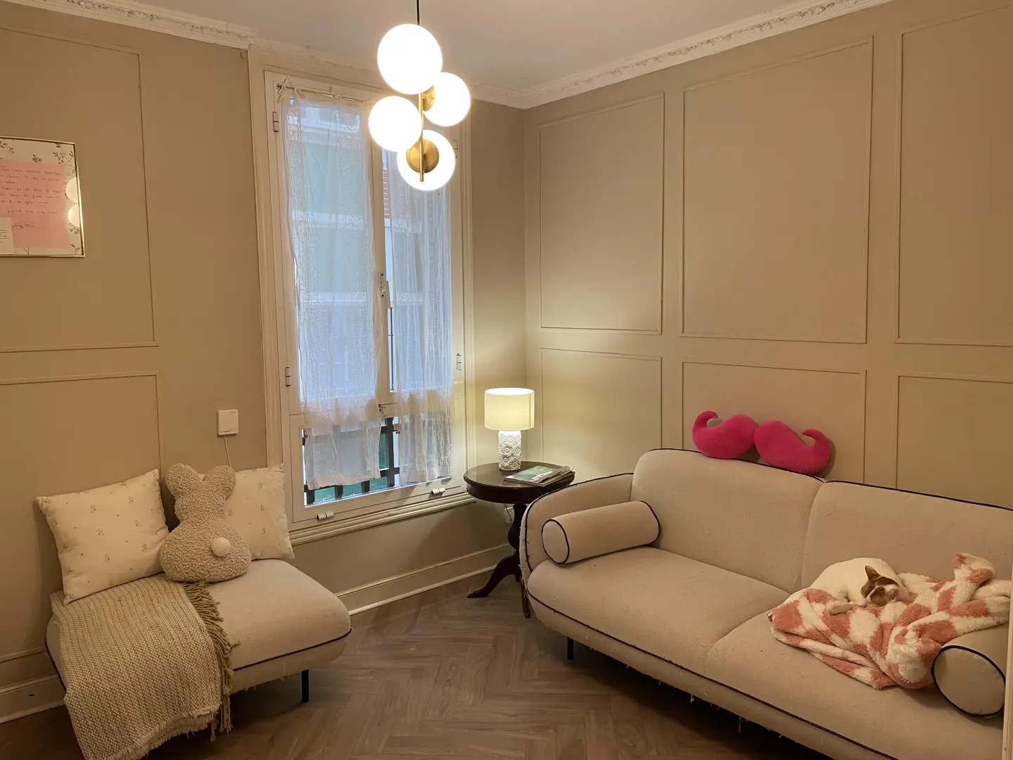 Cozy living room with beige walls, herringbone floor, and white sofa with a dog sleeping on a pink blanket. A bunny pillow sits on a chair near a window.