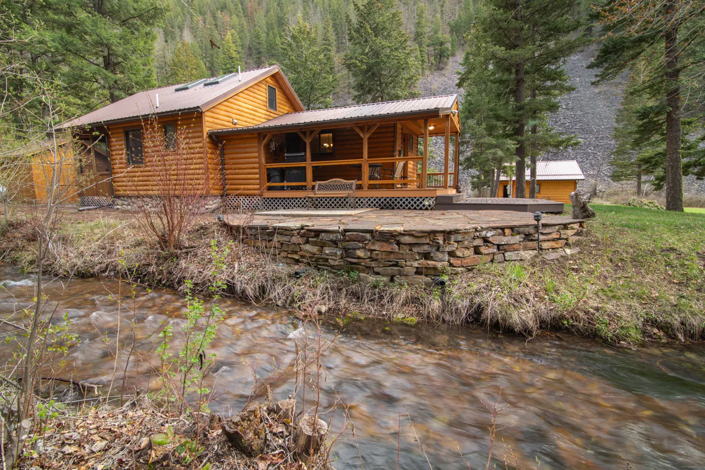 Log cabin with a brown roof and porch sits above a stone wall and flowing creek in a forest setting.
