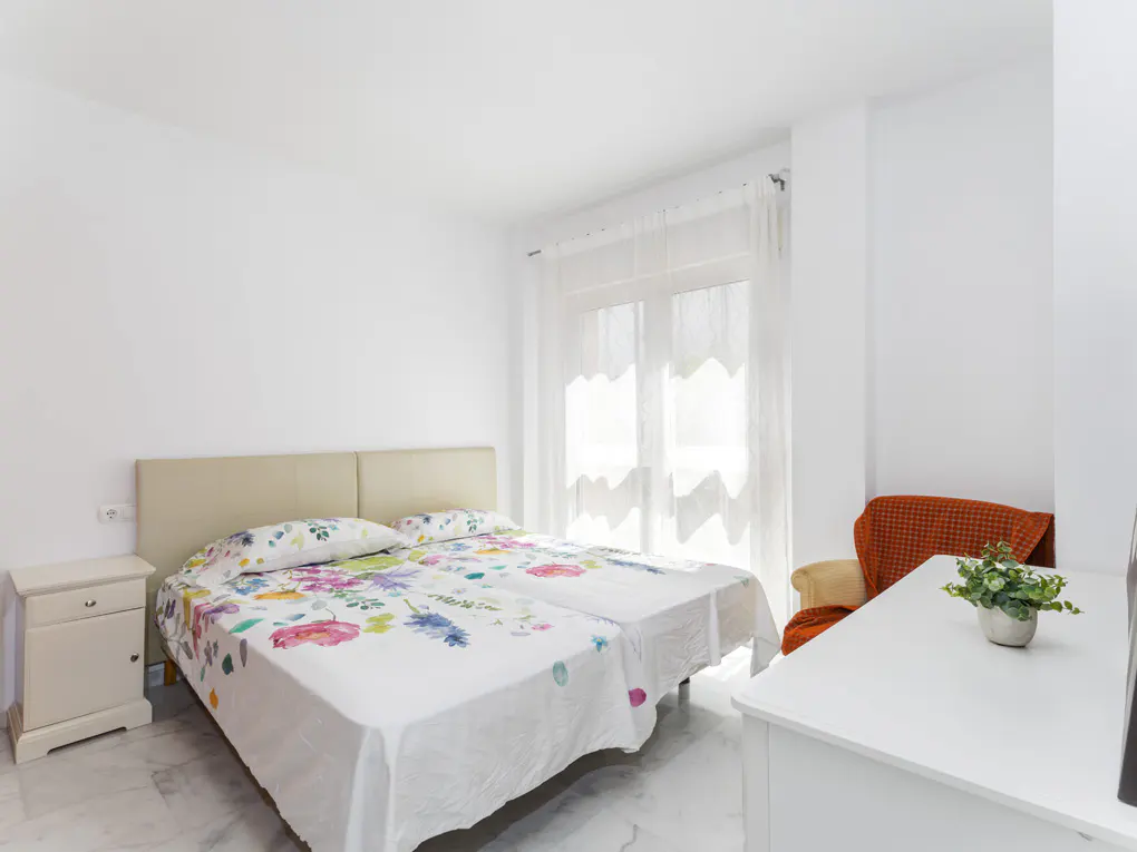 Bright bedroom with two twin beds, floral bedding, white walls, and a white dresser with a plant. A window with sheer curtains lets in natural light.