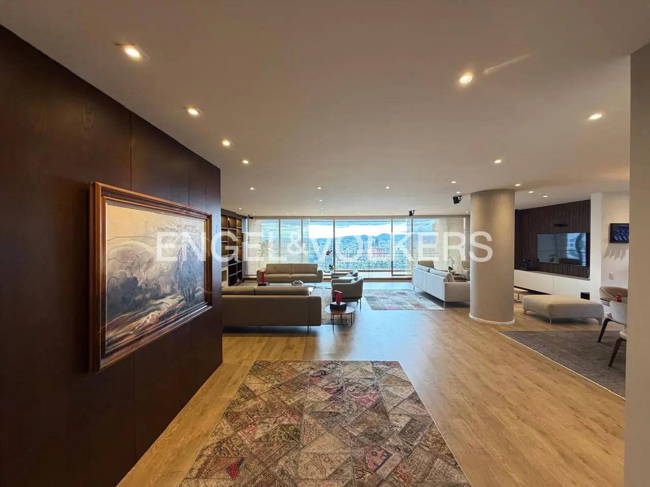 Wide shot of a modern living room with wood floors, sofas, and a large window with a view. A colorful rug is in the foreground.