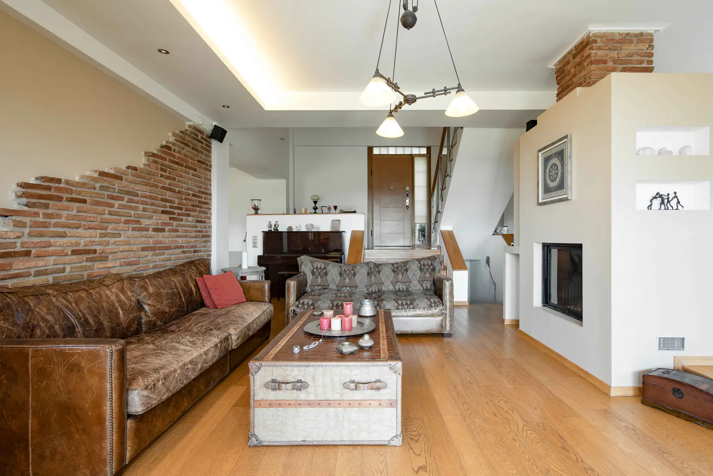 Living room with hardwood floors, leather sofa, patterned loveseat, and trunk-style coffee table. Brick accent wall and fireplace.
