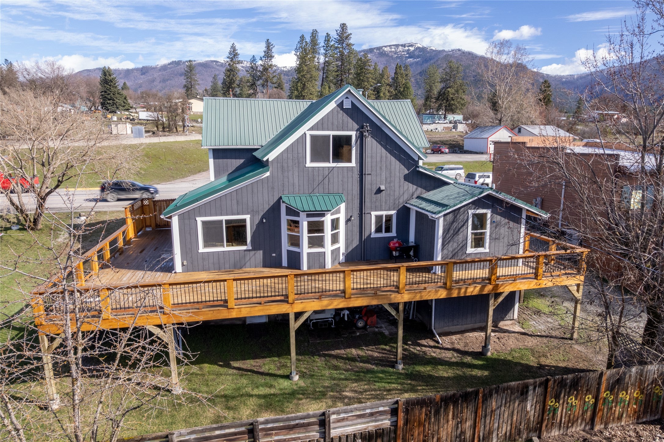 Two-story gray house with a green metal roof and a large wooden deck, set against a backdrop of mountains and trees. [AI-generated text]
