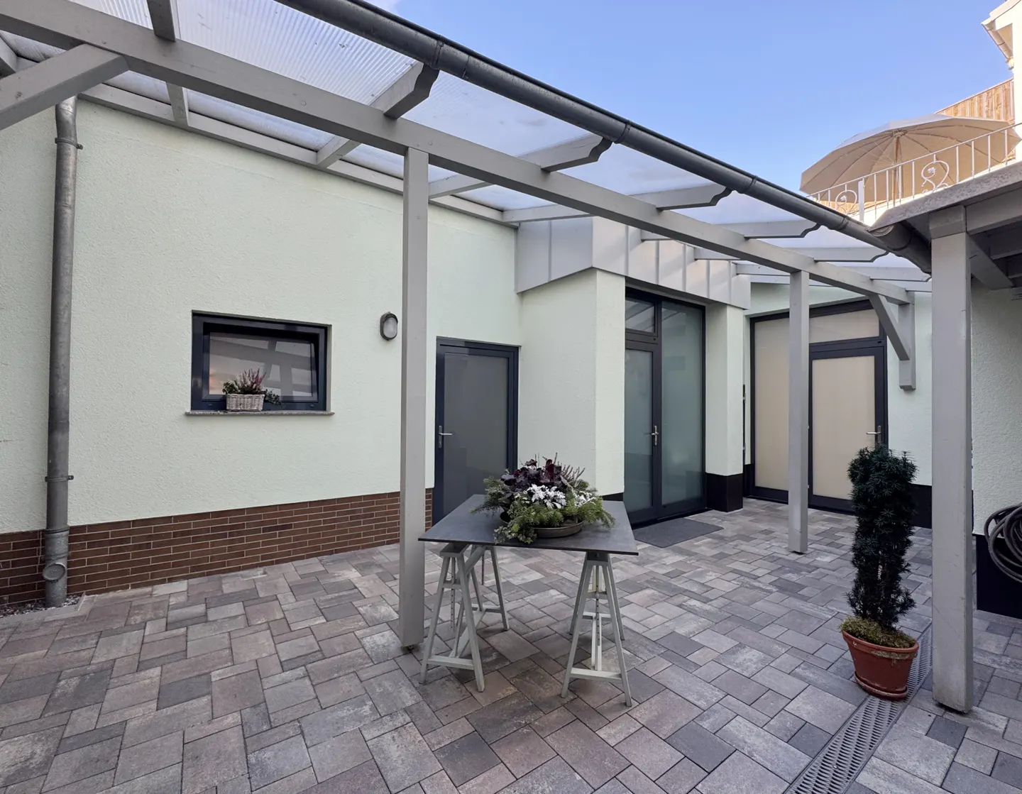 A courtyard with a gray stone patio, a table with a floral arrangement, and a potted plant. The courtyard is covered by a gray pergola.