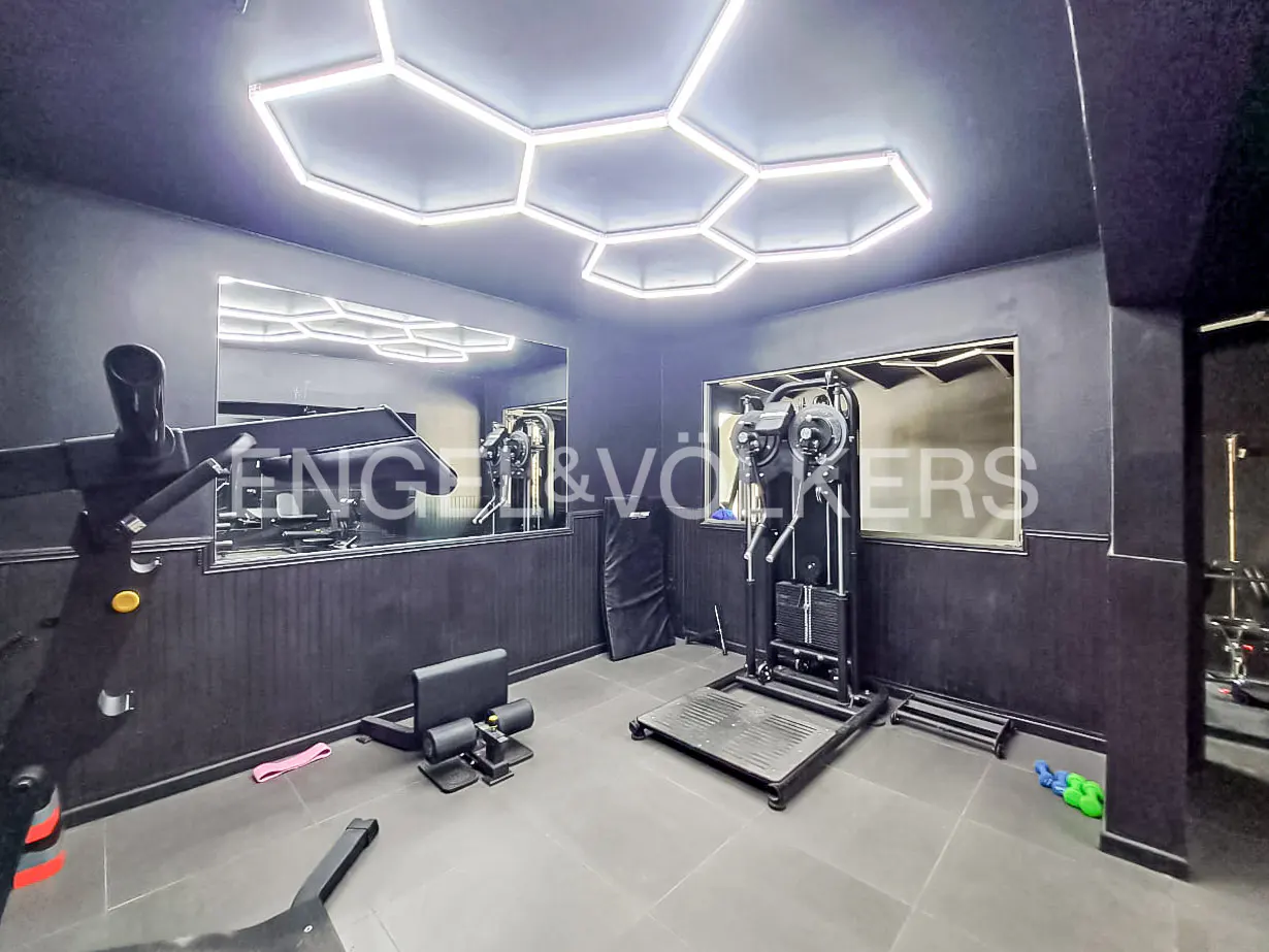 A modern home gym with black walls, gray floors, and hexagon LED ceiling lights. Exercise equipment includes a squat machine and leg press.