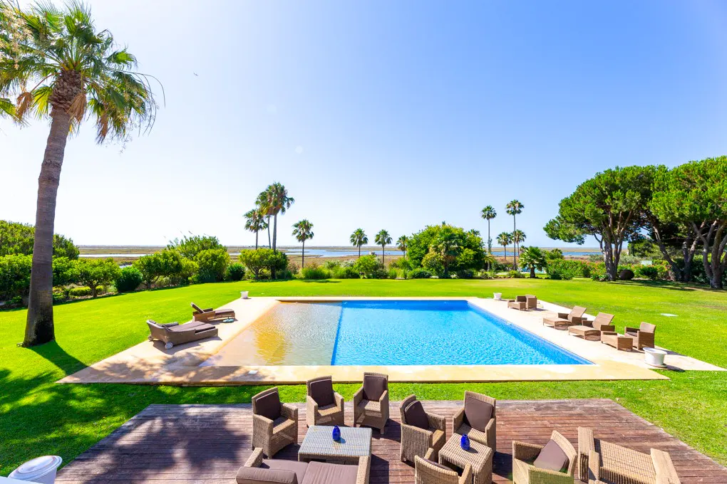 Outdoor pool with lounge chairs, palm trees, and green lawn under a clear blue sky. Wicker furniture on a wooden deck.