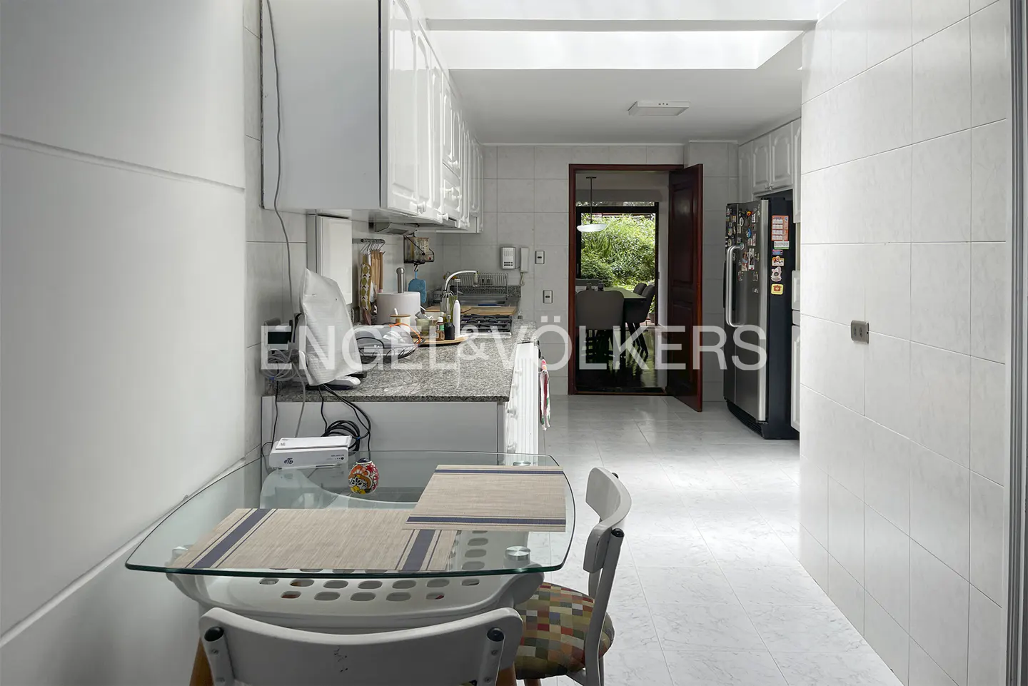 Bright kitchen with white cabinets, tile floors, and a glass table with placemats. A doorway leads to a dining area and garden view.