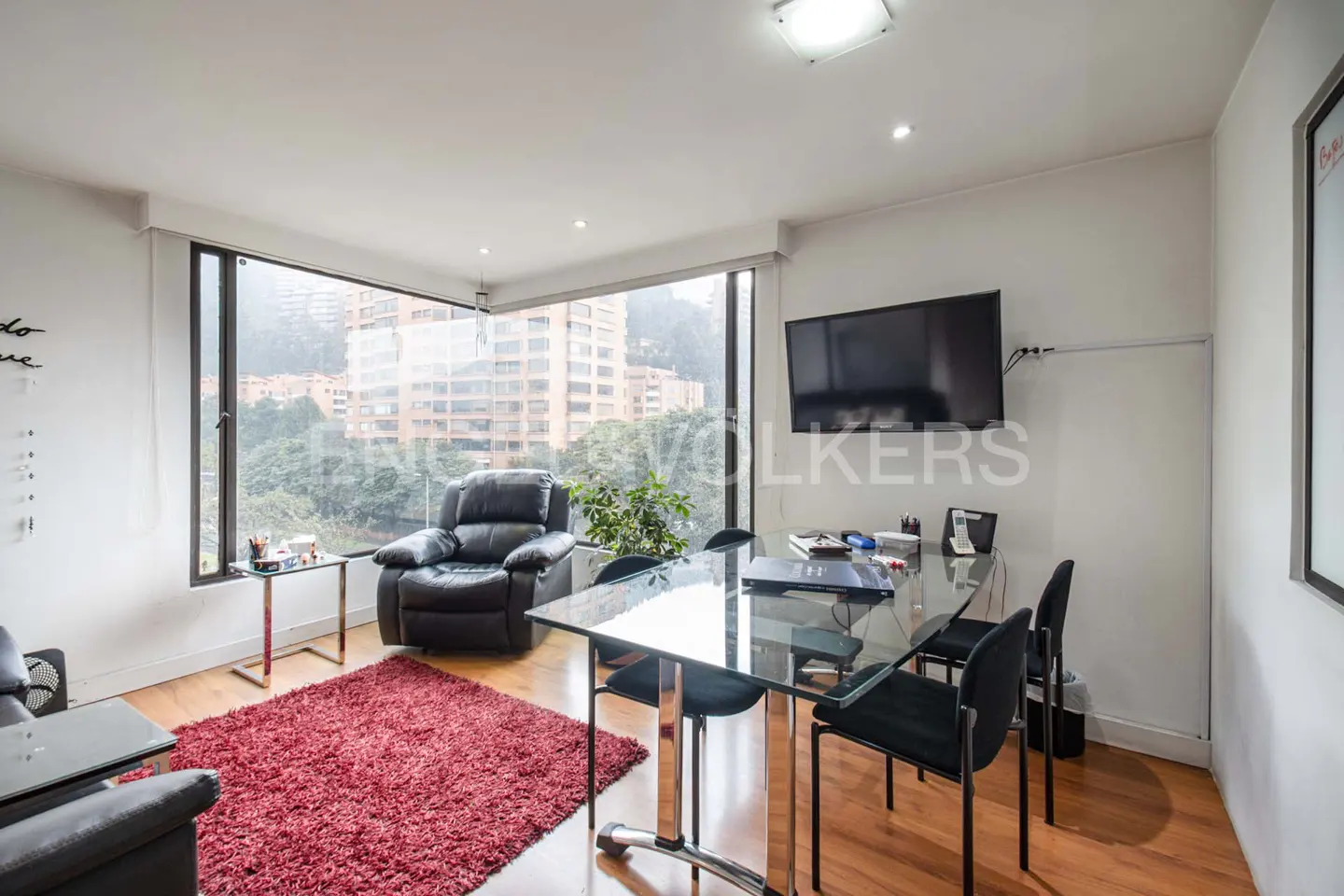 Bright office space with a glass table, black chairs, and a leather recliner near a large window overlooking a cityscape. A red rug adds warmth to the wooden floor.