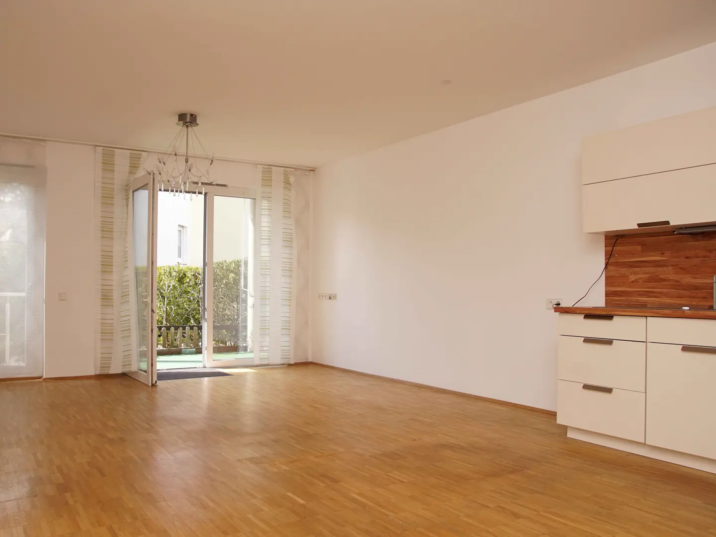 Bright, empty room with wood floors, white walls, and kitchen cabinets. Glass doors lead to a green outdoor space.