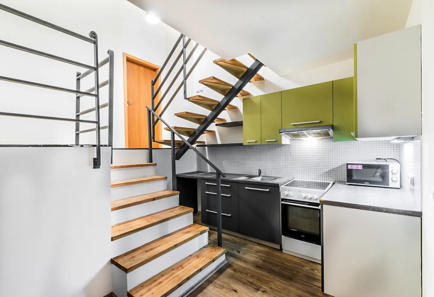 Modern kitchen with olive green and black cabinets, white tiled backsplash, and wooden stairs with a black metal railing.