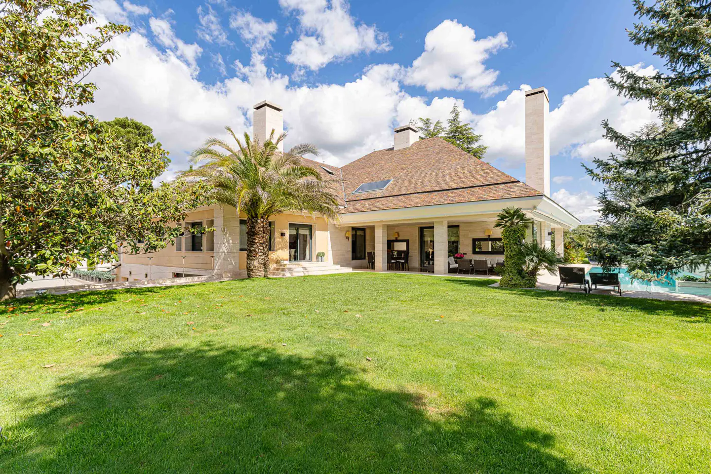 Exterior view of a tan house with a brown roof, palm tree, green lawn, and blue sky with white clouds.