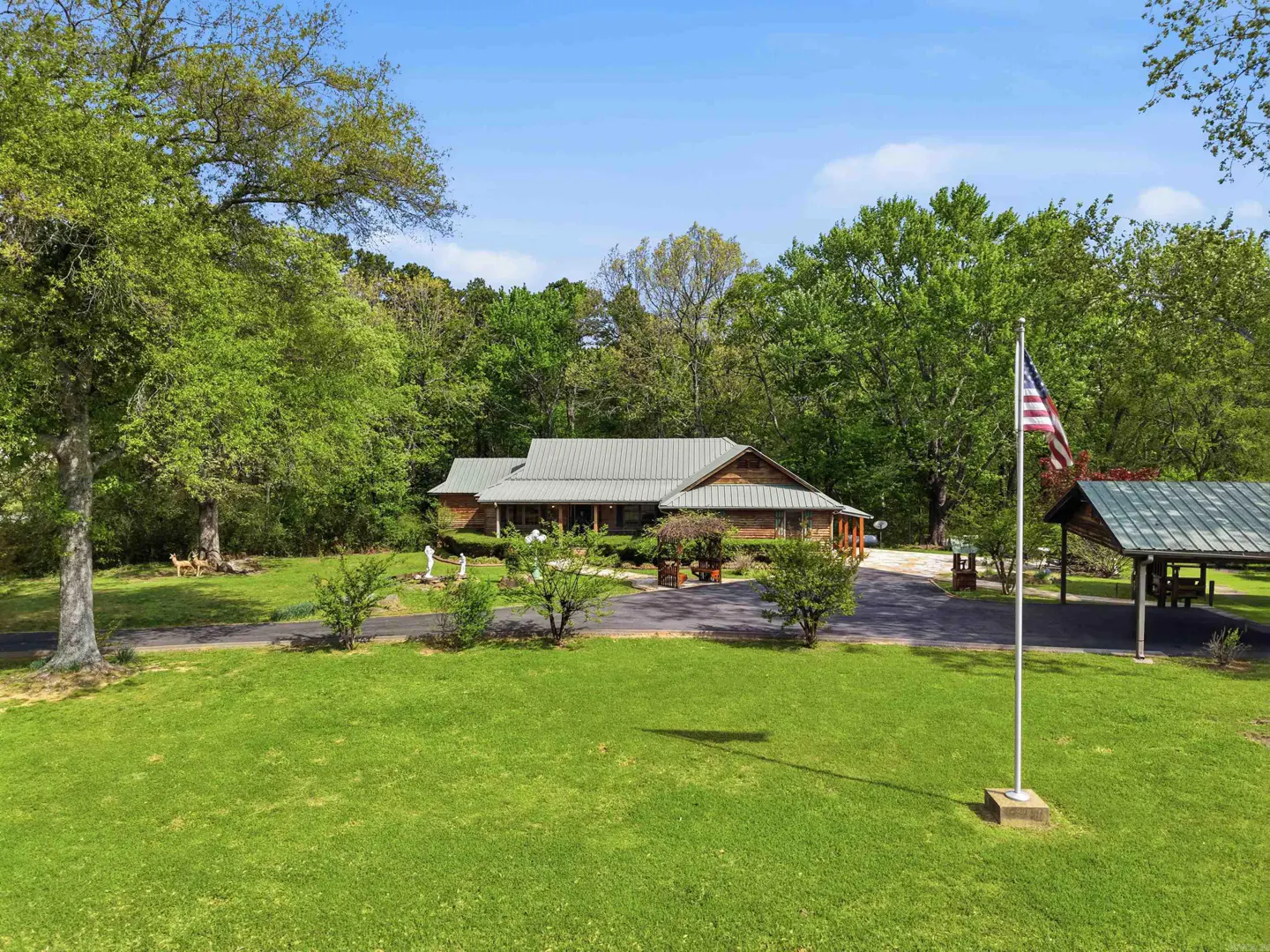 A single-story log cabin with a metal roof, surrounded by green trees and a lawn. An American flag stands tall in the yard.