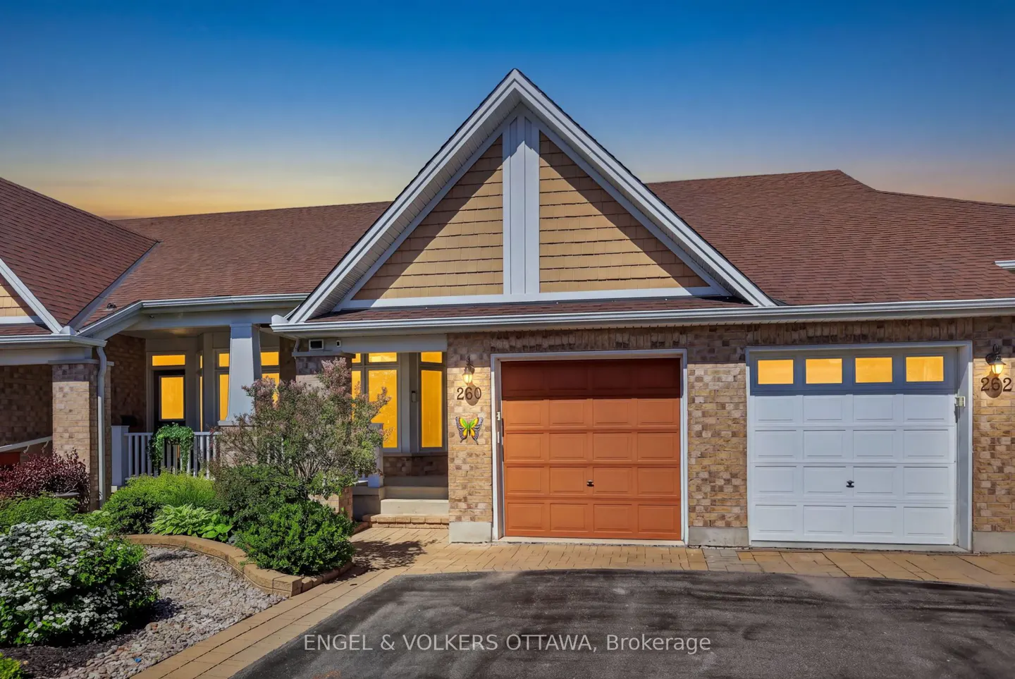 Exterior of a brick townhouse with a brown roof, orange and white garage doors, and a landscaped front yard at sunset.