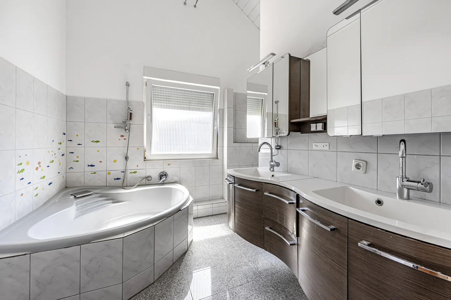 Modern bathroom with a corner bathtub, double vanity with wood cabinets, white tiled walls, and a window.