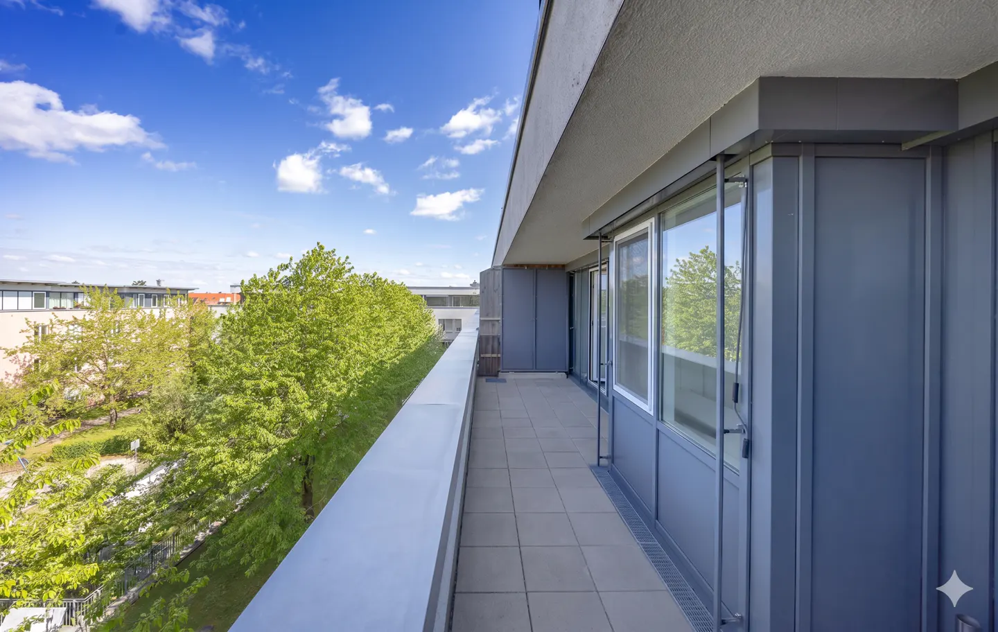 A long, gray balcony with tile flooring overlooks green trees under a blue, cloudy sky.