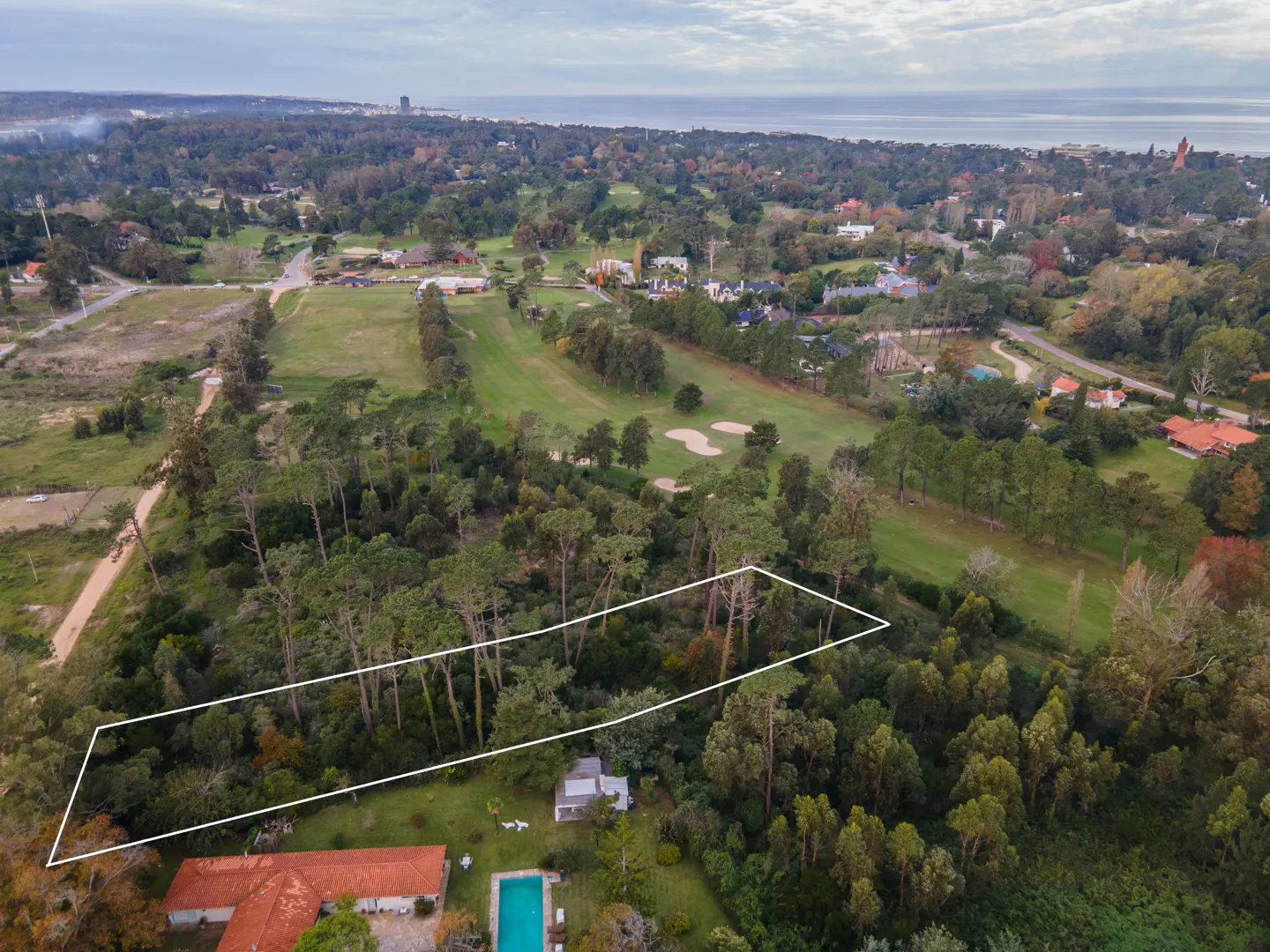 Aerial view of a wooded property outlined in white, with a red-roofed house and pool. Golf course and ocean in the background.