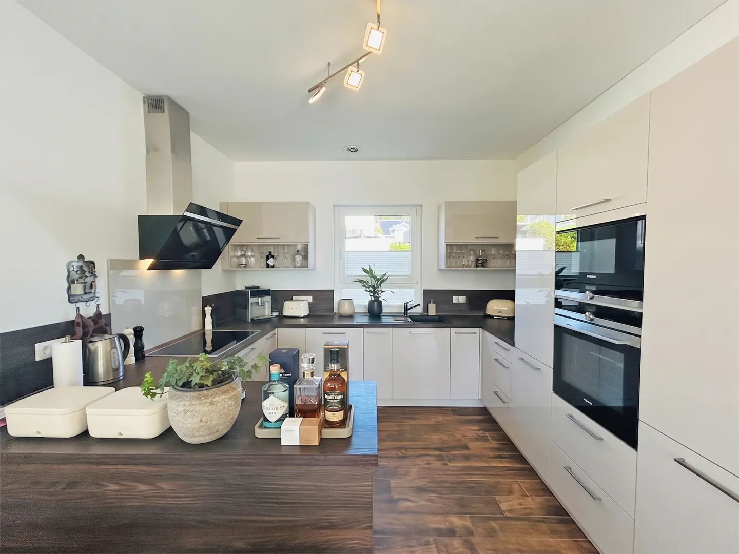 A modern kitchen with white cabinets, dark countertops, and wood floors. Appliances include a black range hood and built-in ovens.