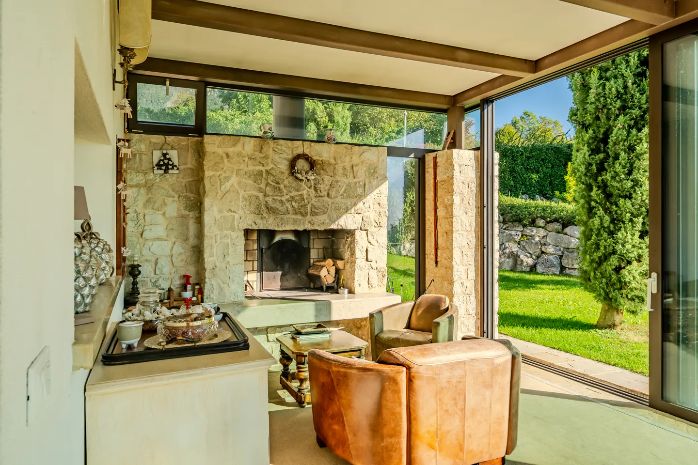 Living room with stone fireplace, brown leather chairs, and sliding glass doors to a green lawn and stone wall.