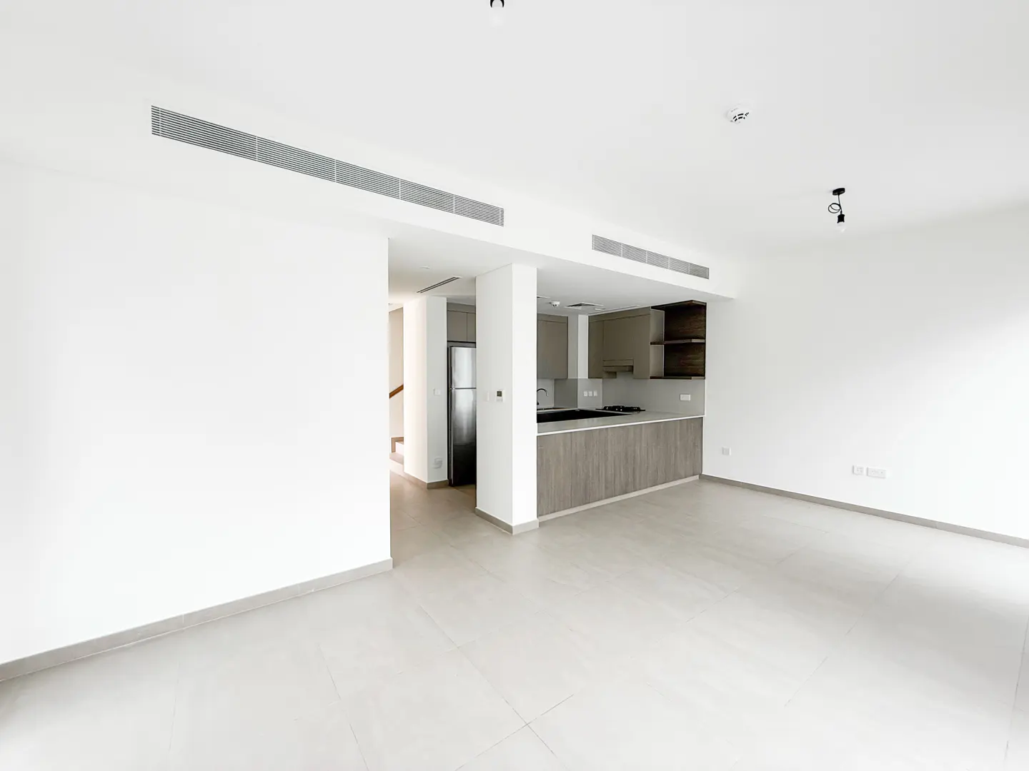 Bright, empty living room with white walls and tile floor. An open kitchen with gray cabinets is visible in the background.