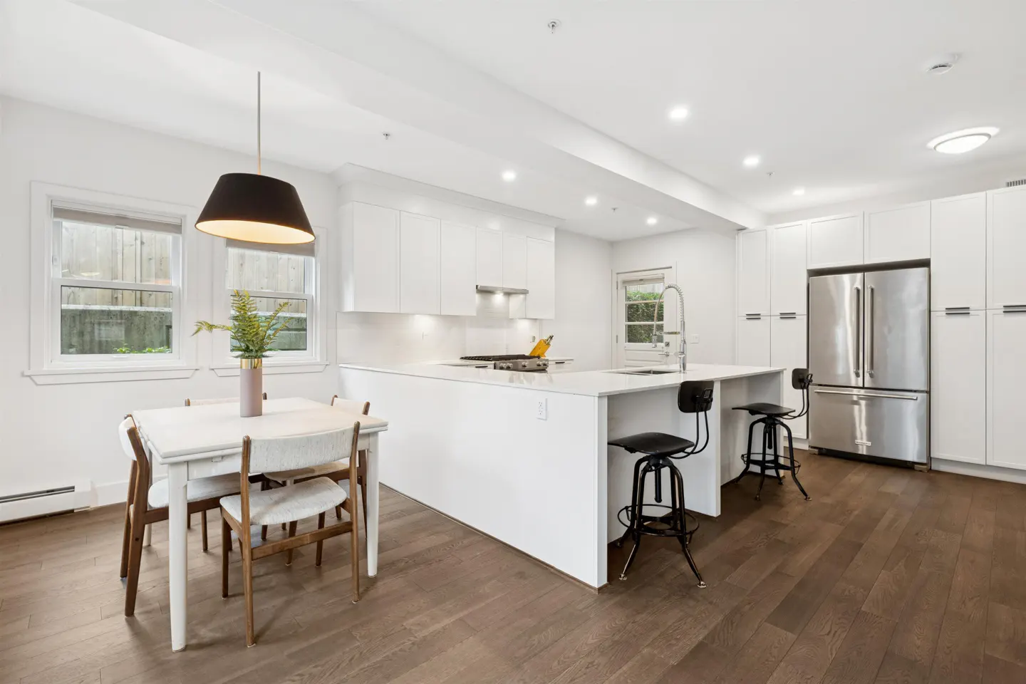 Bright, modern kitchen with white cabinets, stainless steel fridge, island with stools, and a dining table with a black pendant light.