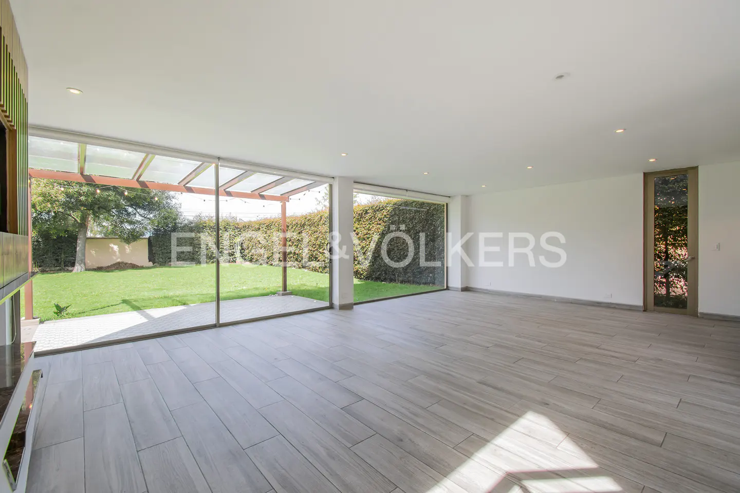 Modern living room with light wood-look tile floors, floor-to-ceiling glass walls, and a view of a lush green backyard.