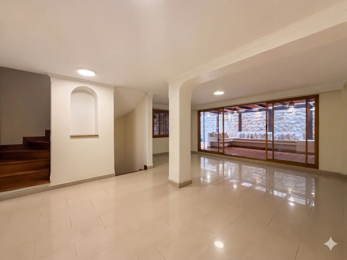 Bright, open-plan living area with beige tile flooring, white walls, and wooden stairs. Sliding glass doors lead to an outdoor seating area.