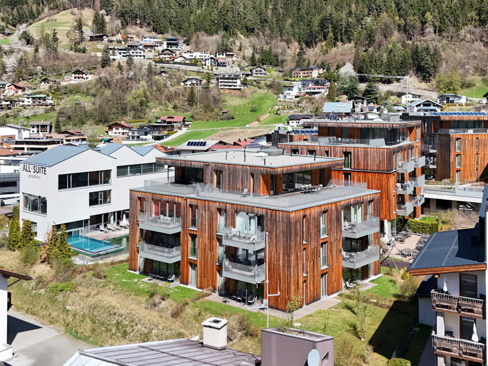 Exterior view of modern wooden apartments with balconies, near a white resort building and a pool, set against a hillside village and forest. [AI-generated text]