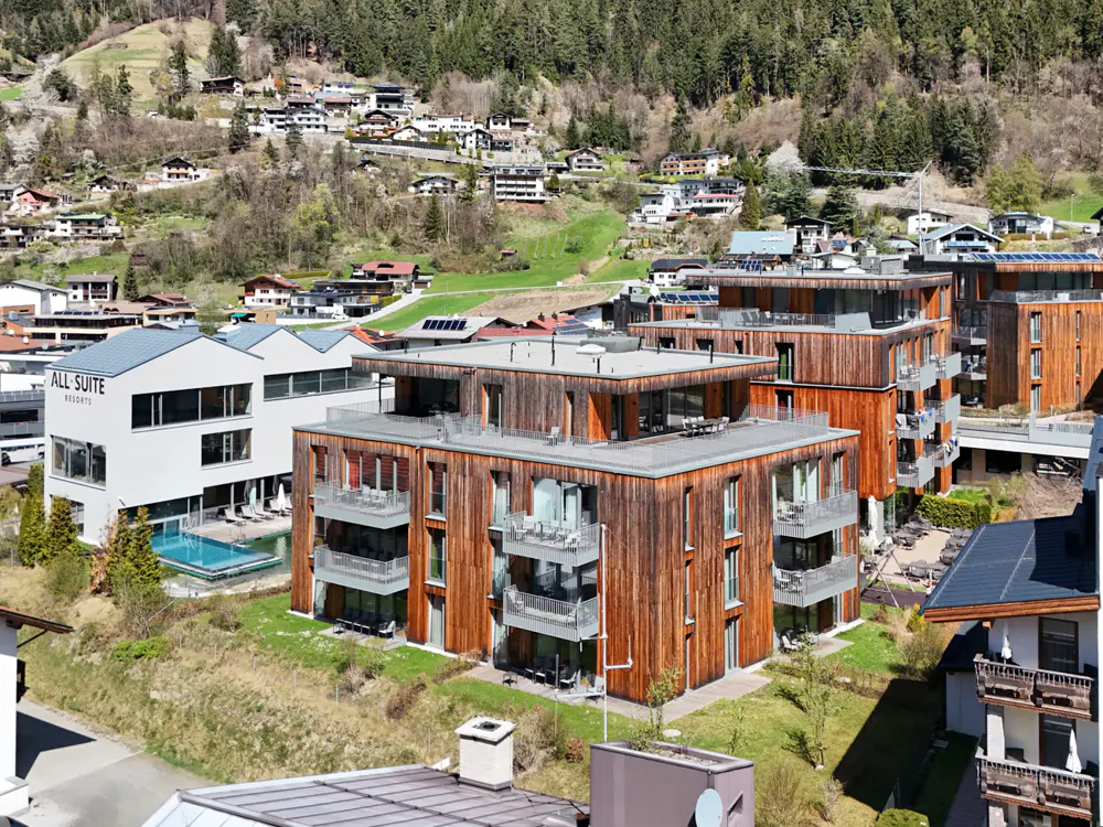 Exterior view of modern wooden apartments with balconies, near a white resort building and a pool, set against a hillside village and forest.