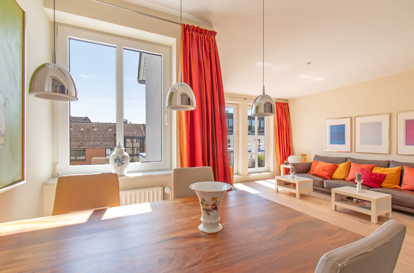 Bright living and dining room with a wooden table, grey sofa, vibrant orange curtains, and modern pendant lighting.
