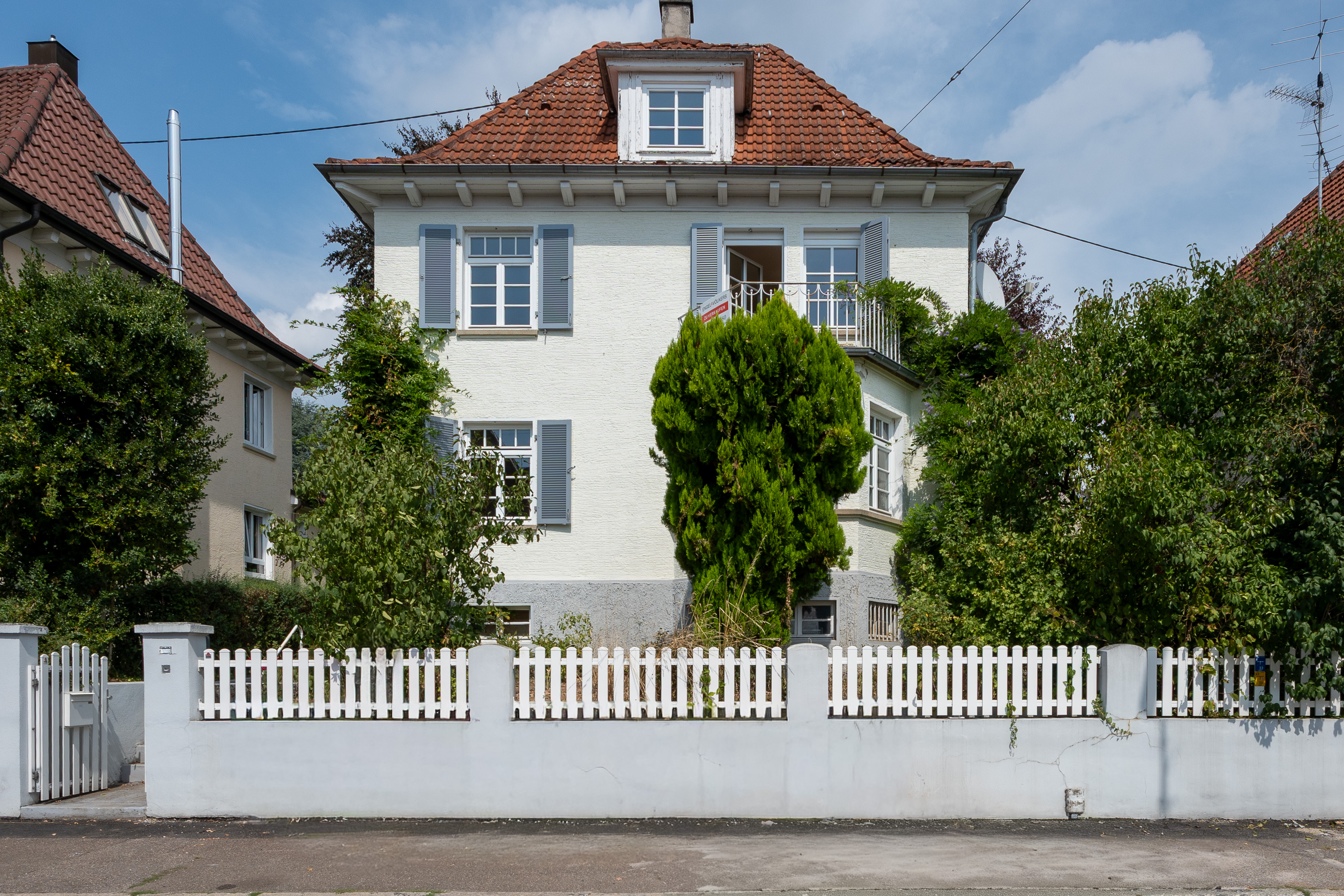 A cream-colored house with a red tile roof, blue shutters, and a white picket fence, surrounded by lush green trees. [AI-generated text]