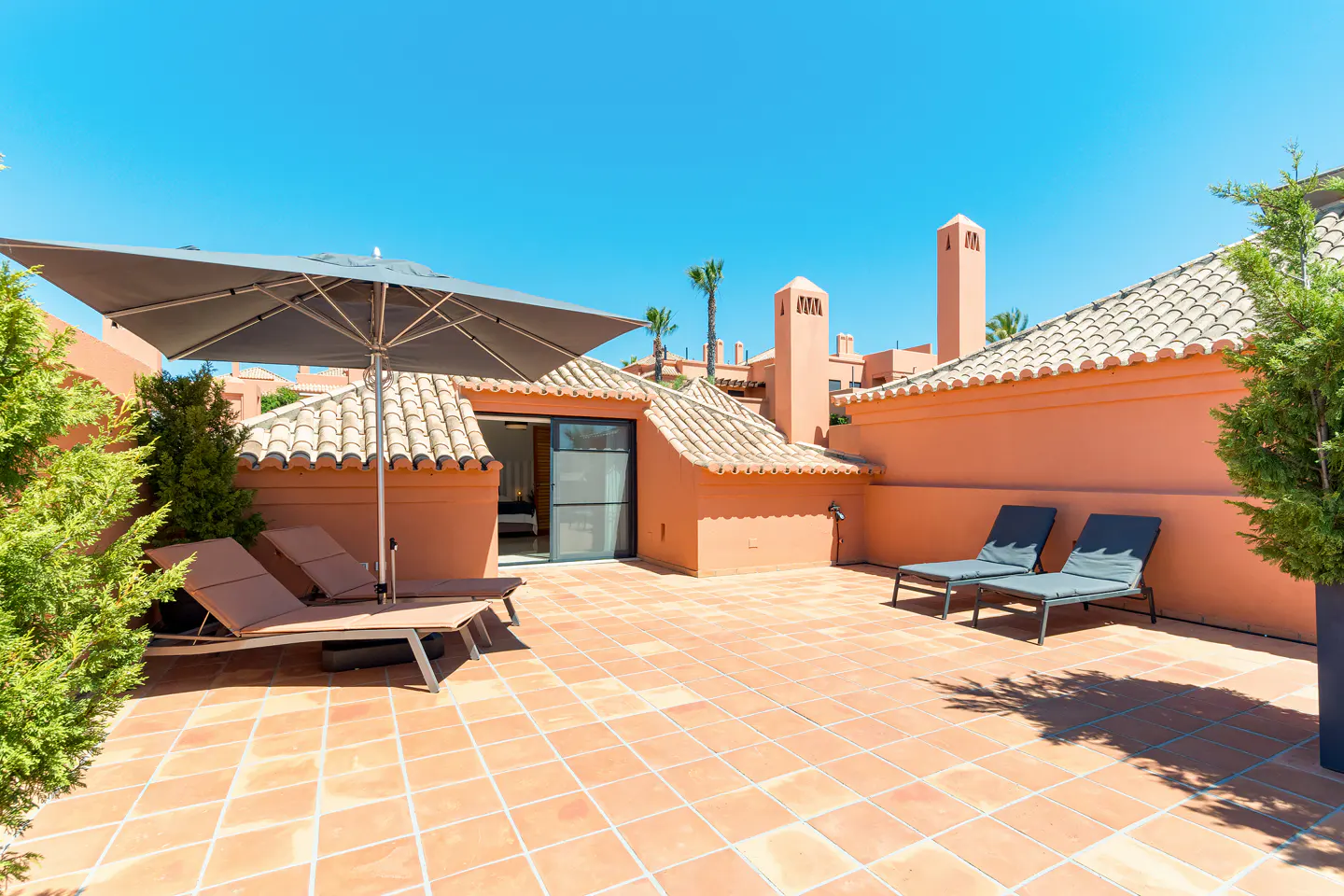 Terrace view with terracotta tiles, peach walls, and blue sky. Lounge chairs, umbrella, and greenery add to the outdoor living space.