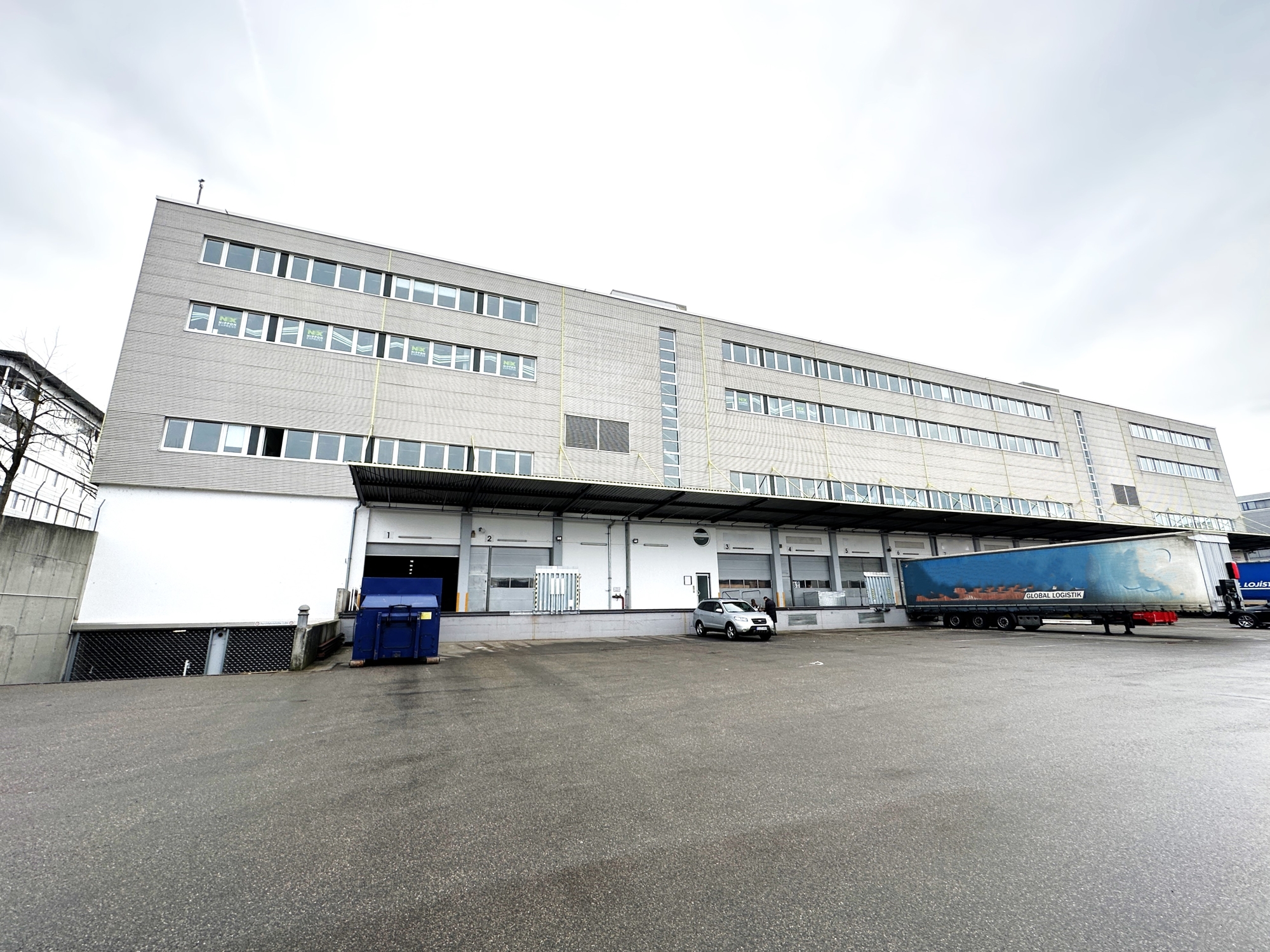 Large industrial warehouse with loading docks, a parked semi-truck, and a silver car in a paved lot under a cloudy sky.