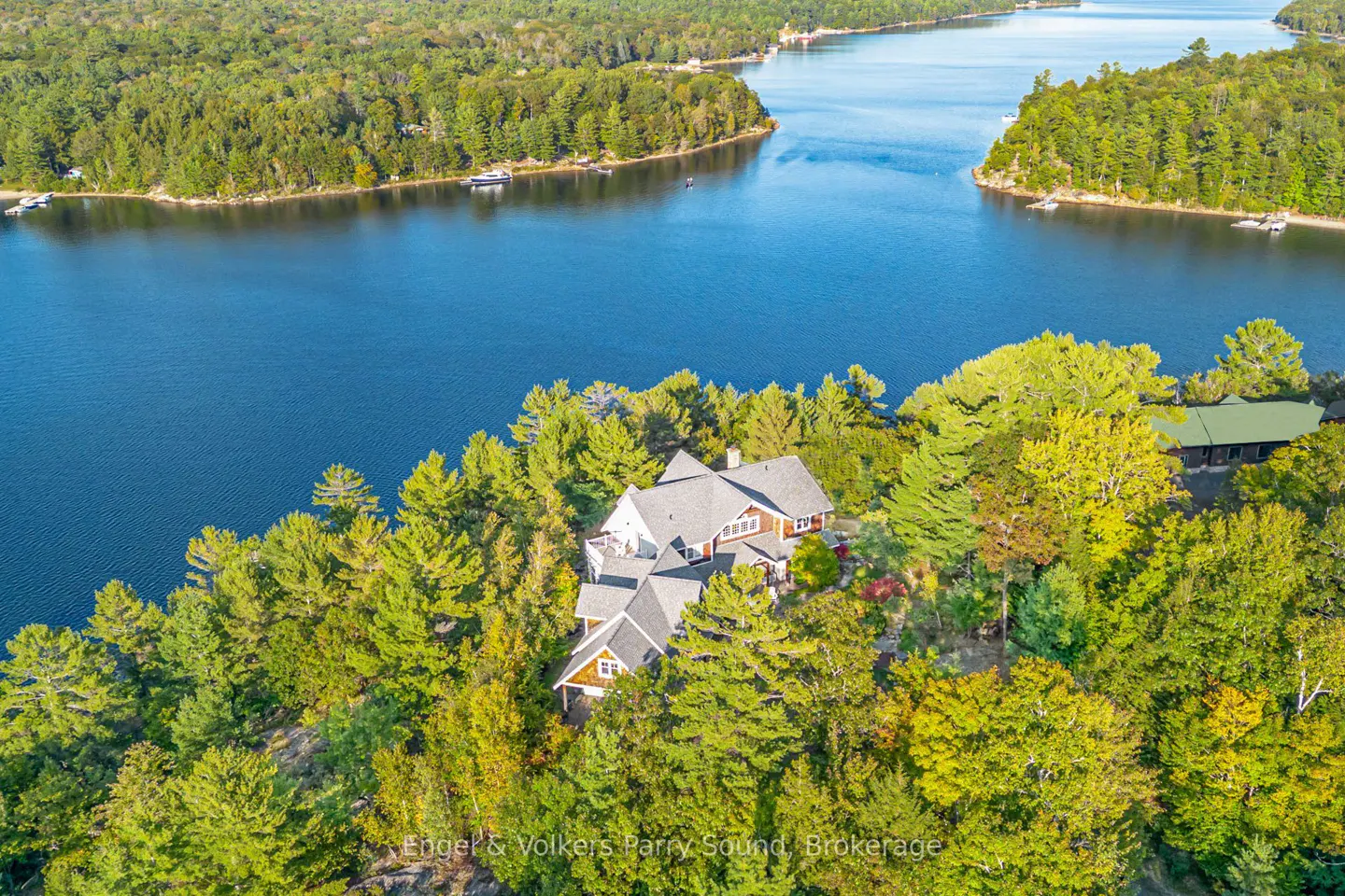 Aerial view of a large, brown house with a gray roof surrounded by green trees, next to a blue lake.
