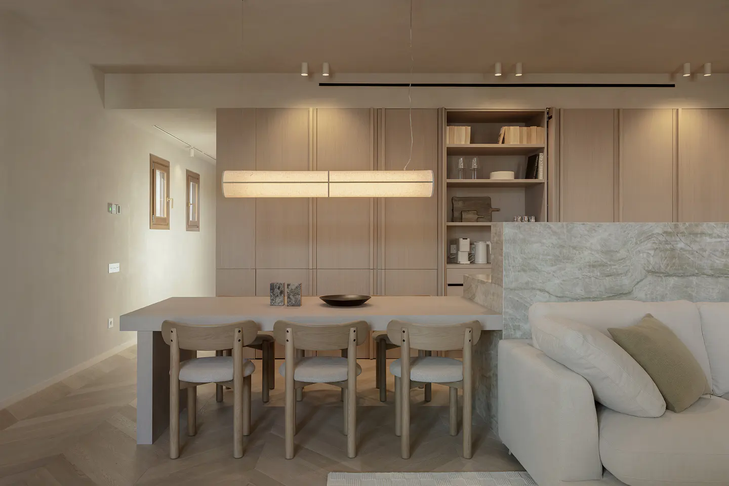 Beige dining room with a long table, wooden chairs, and a rectangular light fixture. A white sofa is visible in the foreground.