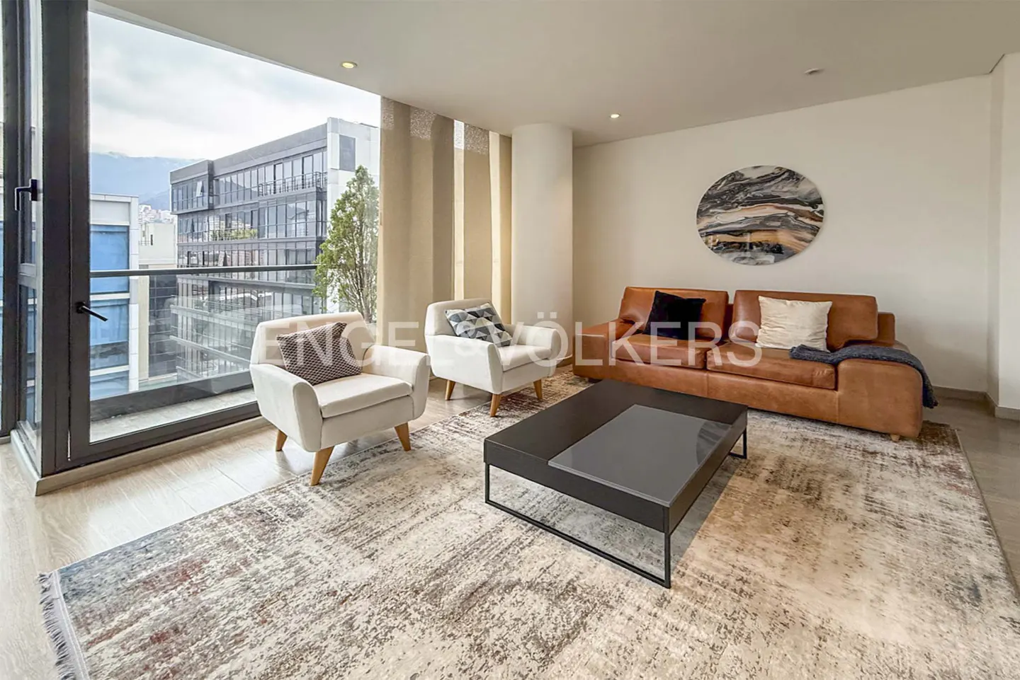 Bright living room with two white armchairs, a brown leather sofa, and a black coffee table on a patterned rug. Balcony with city view.